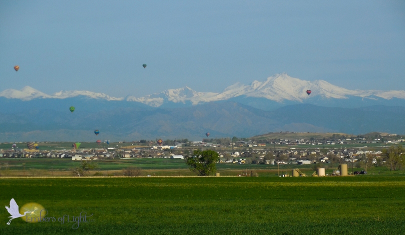 Six brightly colored balloons rise above the plains. Behind them, the Rocky Mountains and Longs Peak are easy to see. The foothills are covered in brush, but the large peaks behind are still covered in snow. A grassy field is in the foreground and urban sprawl between the fields and the foothills. 